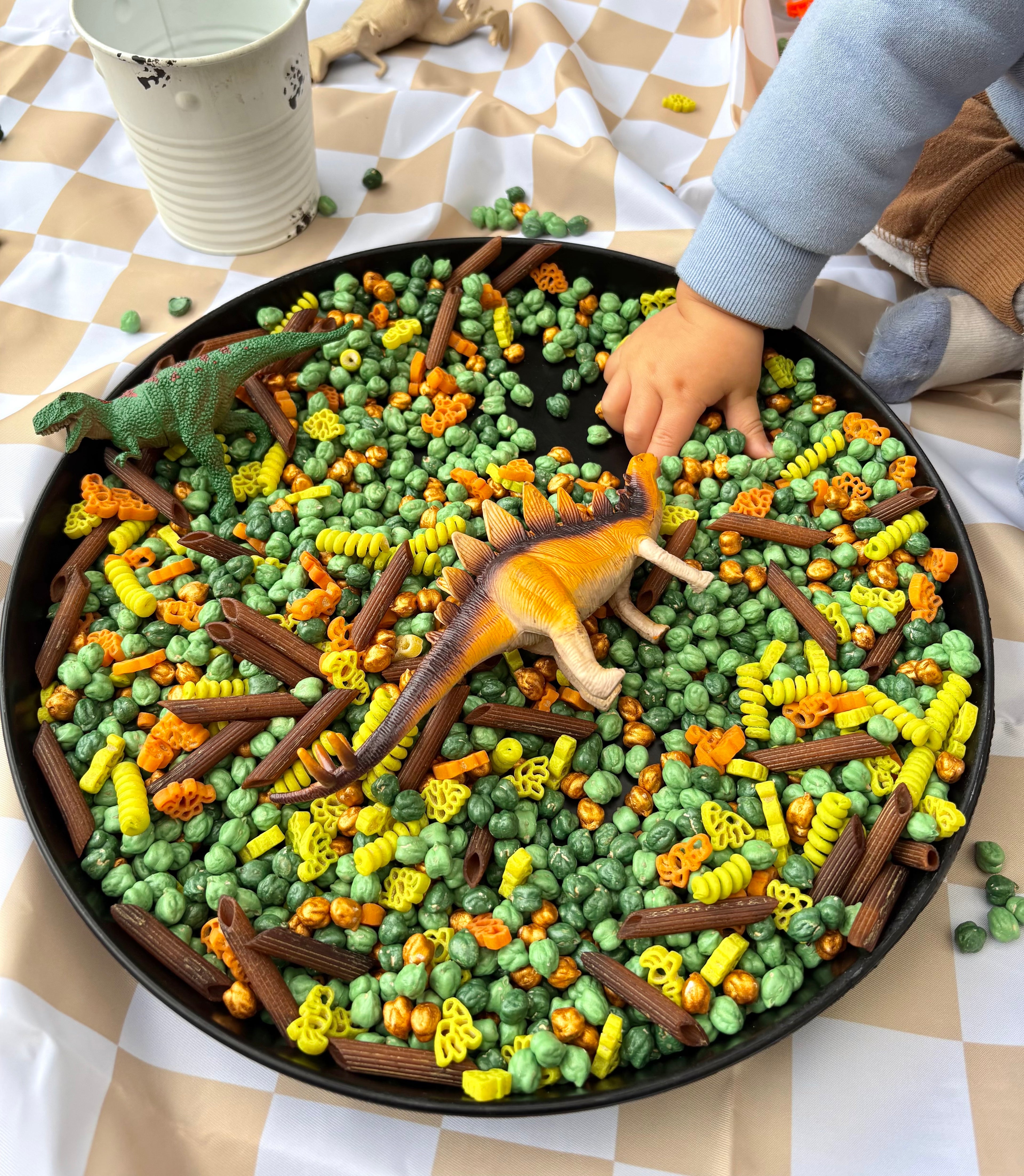 Child playing with colorful pasta and toy dinosaurs in a pan on a checkered tablecloth.