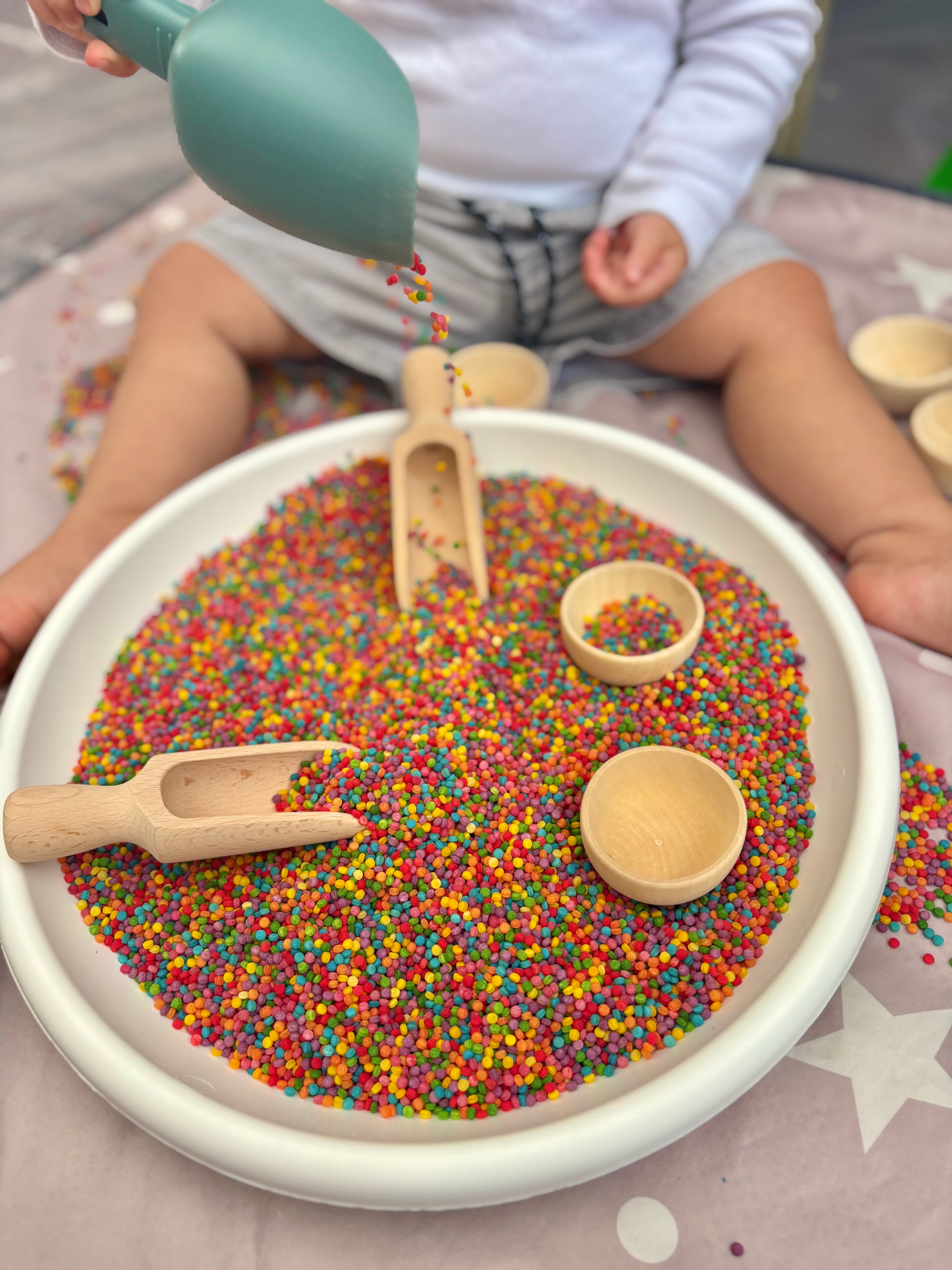 Child playing with colourful sensory bin filler and wooden toys on a white tray.